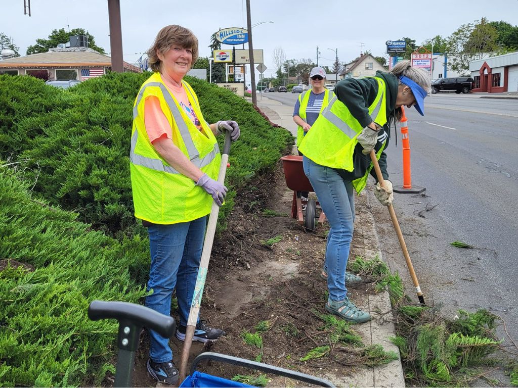 3 people dressed in safety vests who are cleaning brush out of the street. 